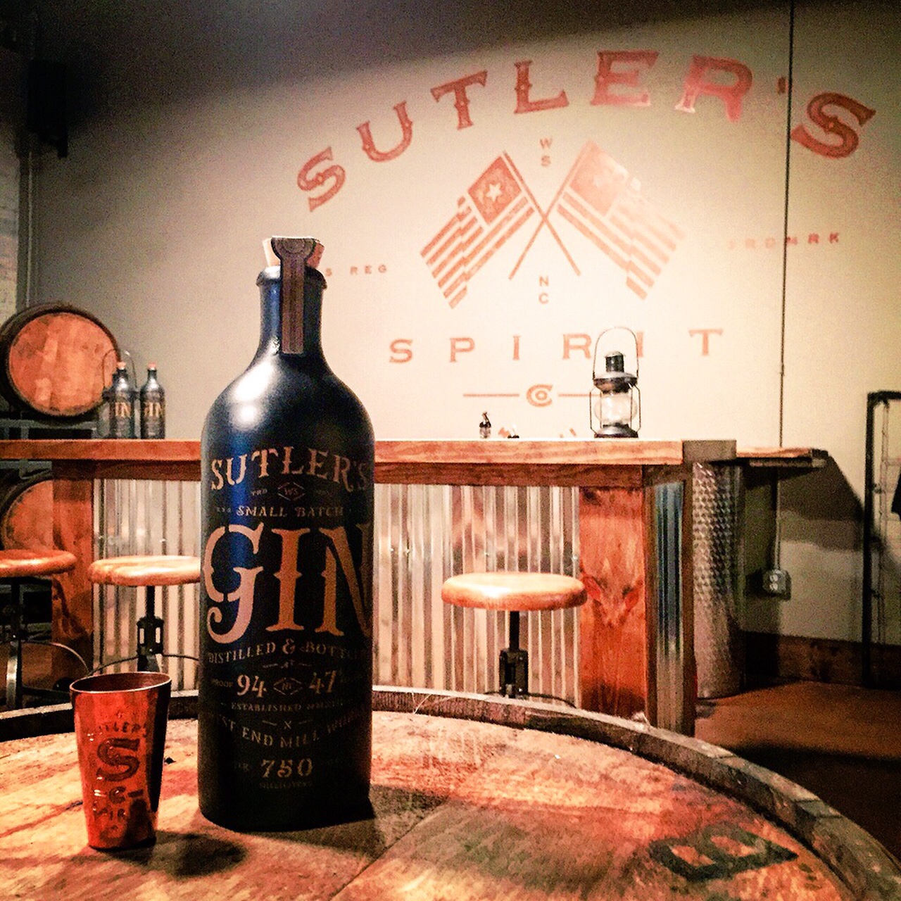 A bottle of gin next to a copper shot glass, both sitting on top of a wooden barrel at a bar