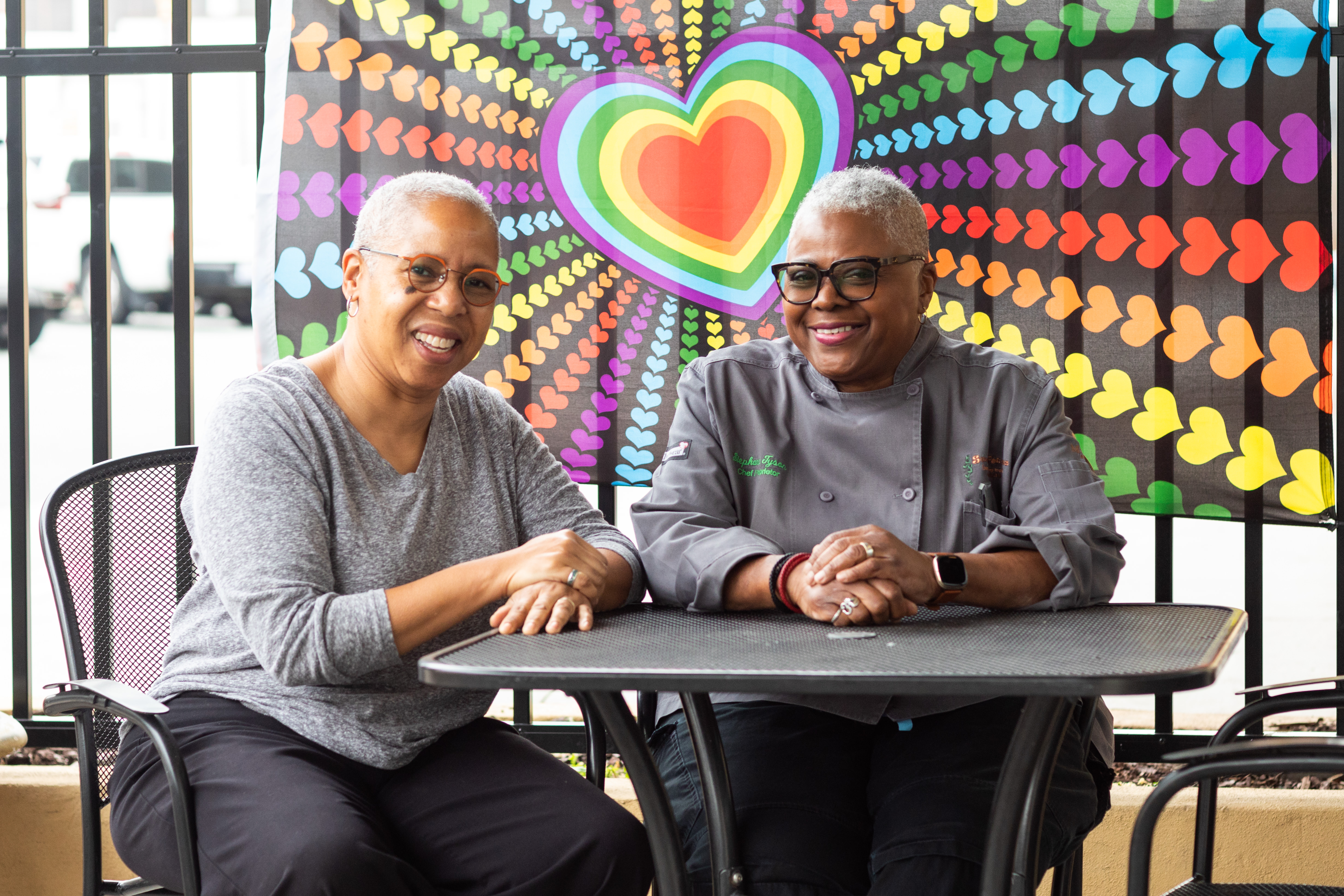 Two women sitting together at a table in front of a rainbow heart flag