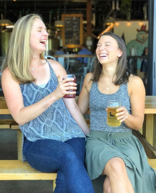 Two women sitting on a bench laughing, holding glasses of kombucha