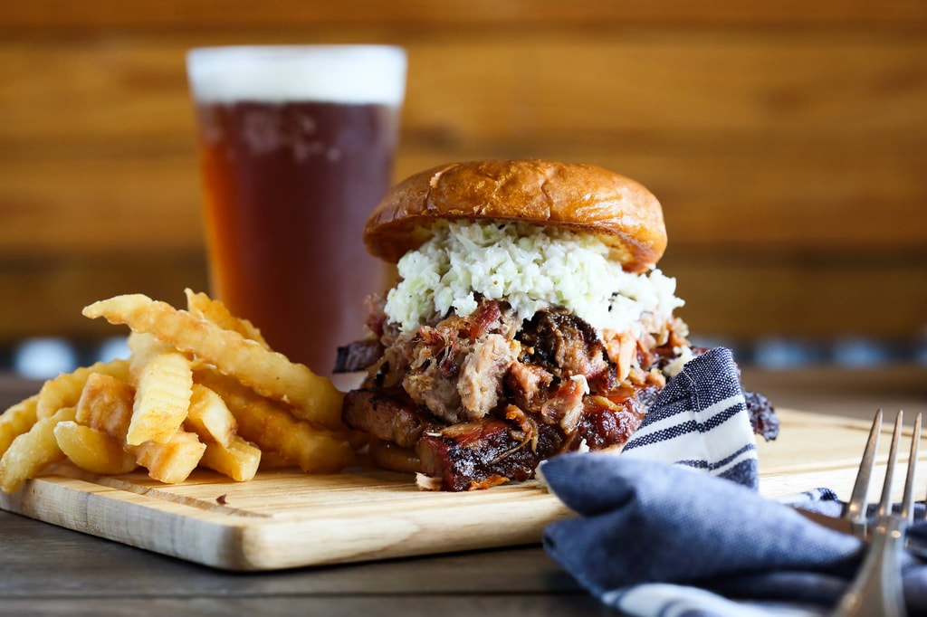 A barbeque pork sandwich with coleslaw on a small wooden cutting board with french fries and an amber-colored beer.