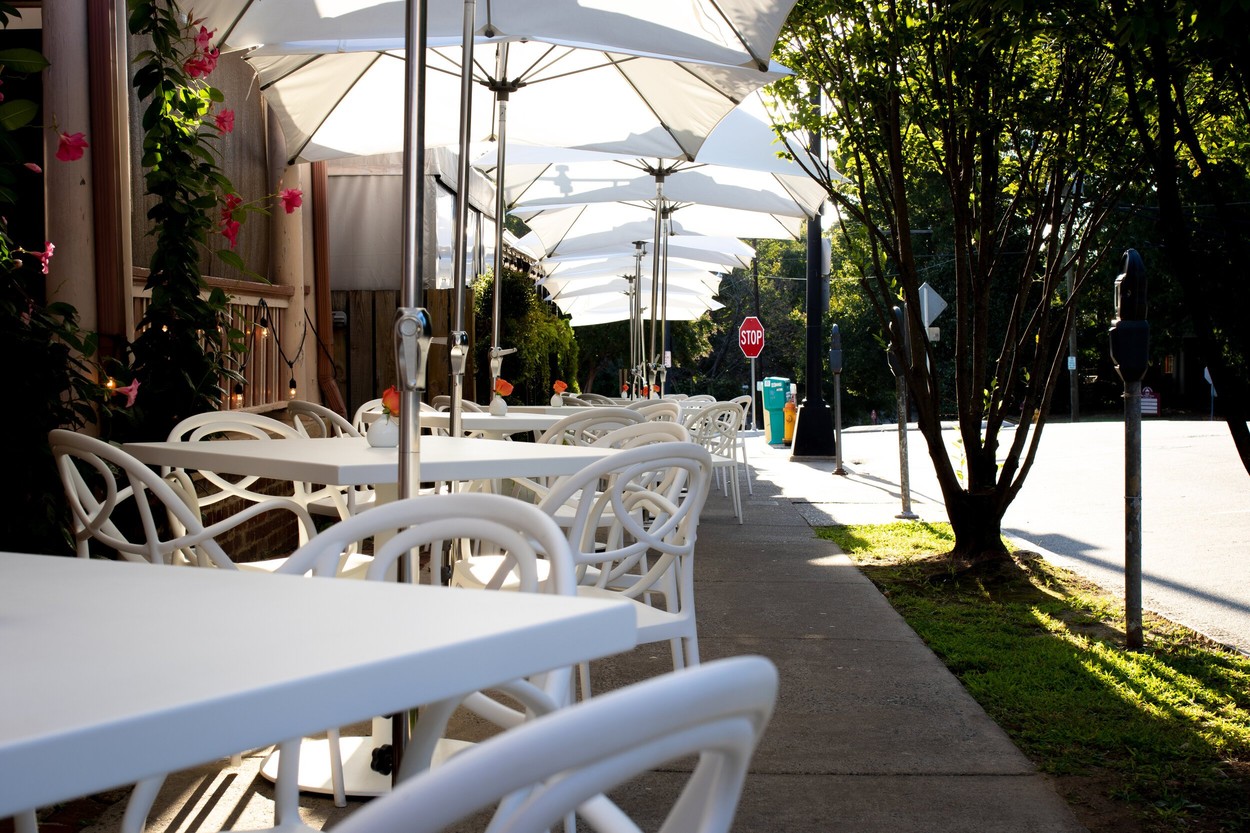 An outdoor dining patio on a tree-lined street with white tables, white chairs, and white umbrellas 