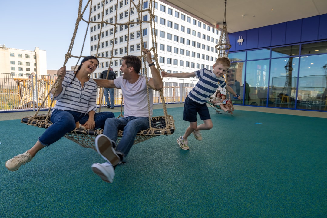 A man and woman sit together on a circular swing on the rooftop of Kaleideum, while a child grabs the swing and twirls them around.