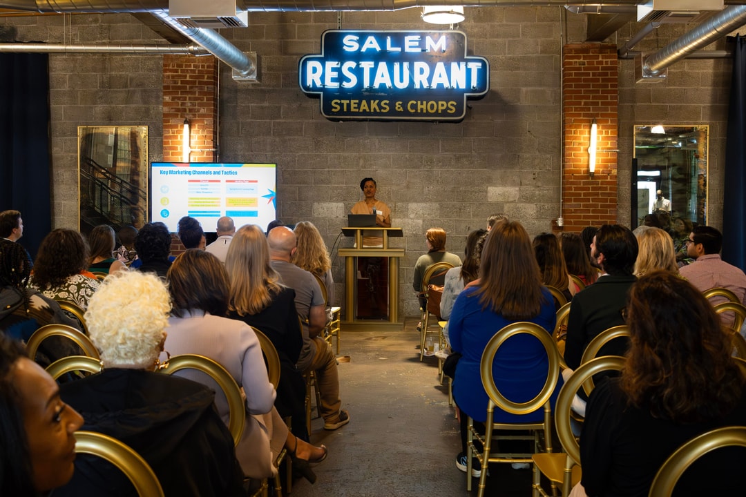 A view from the back of the crowd toward the podium while a speaker showcases a presentation on the main screen at Robert Hall Event Venue.A view from the back of the crowd toward the podium while a speaker showcases a presentation on the main screen at Robert Hall Event Venue.