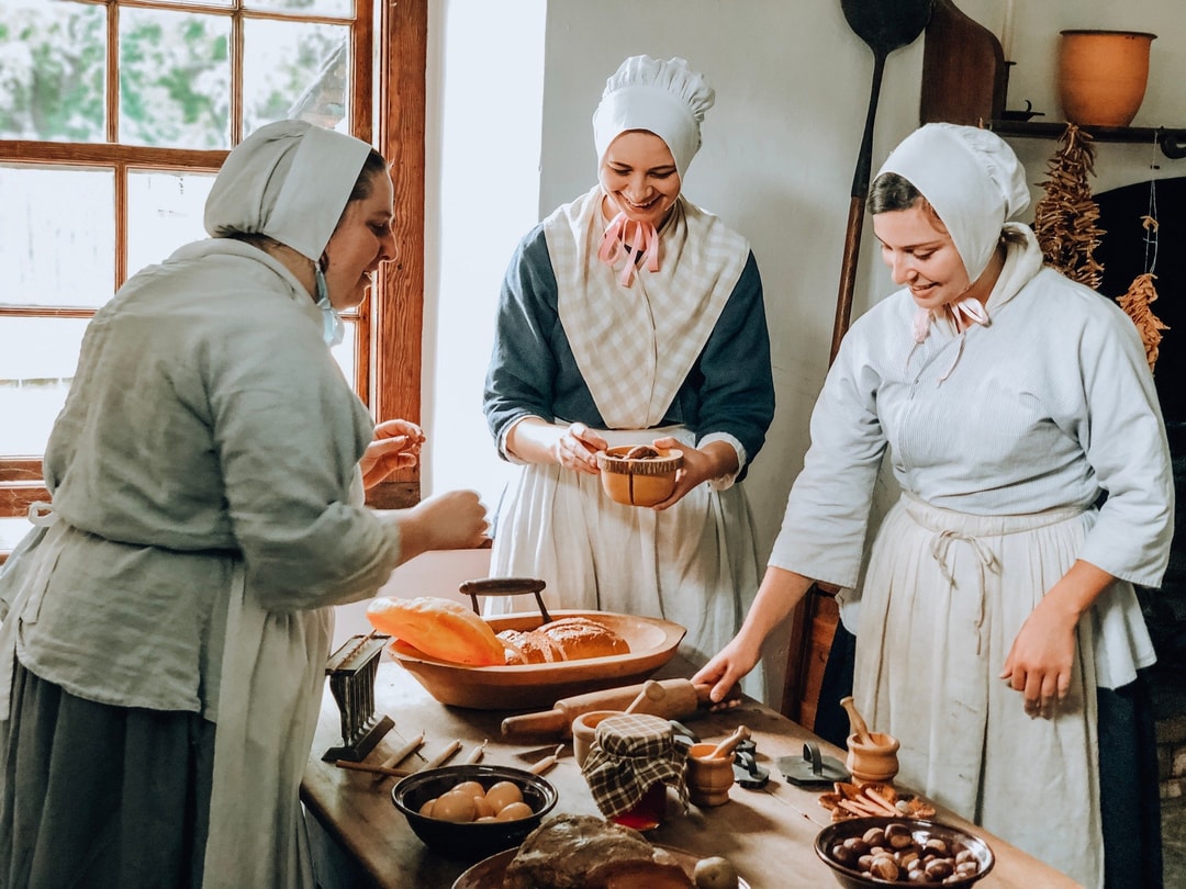Three female costumed interpreters gather around a plate of food at Historic Bethabara Park in traditional Moravian attire.