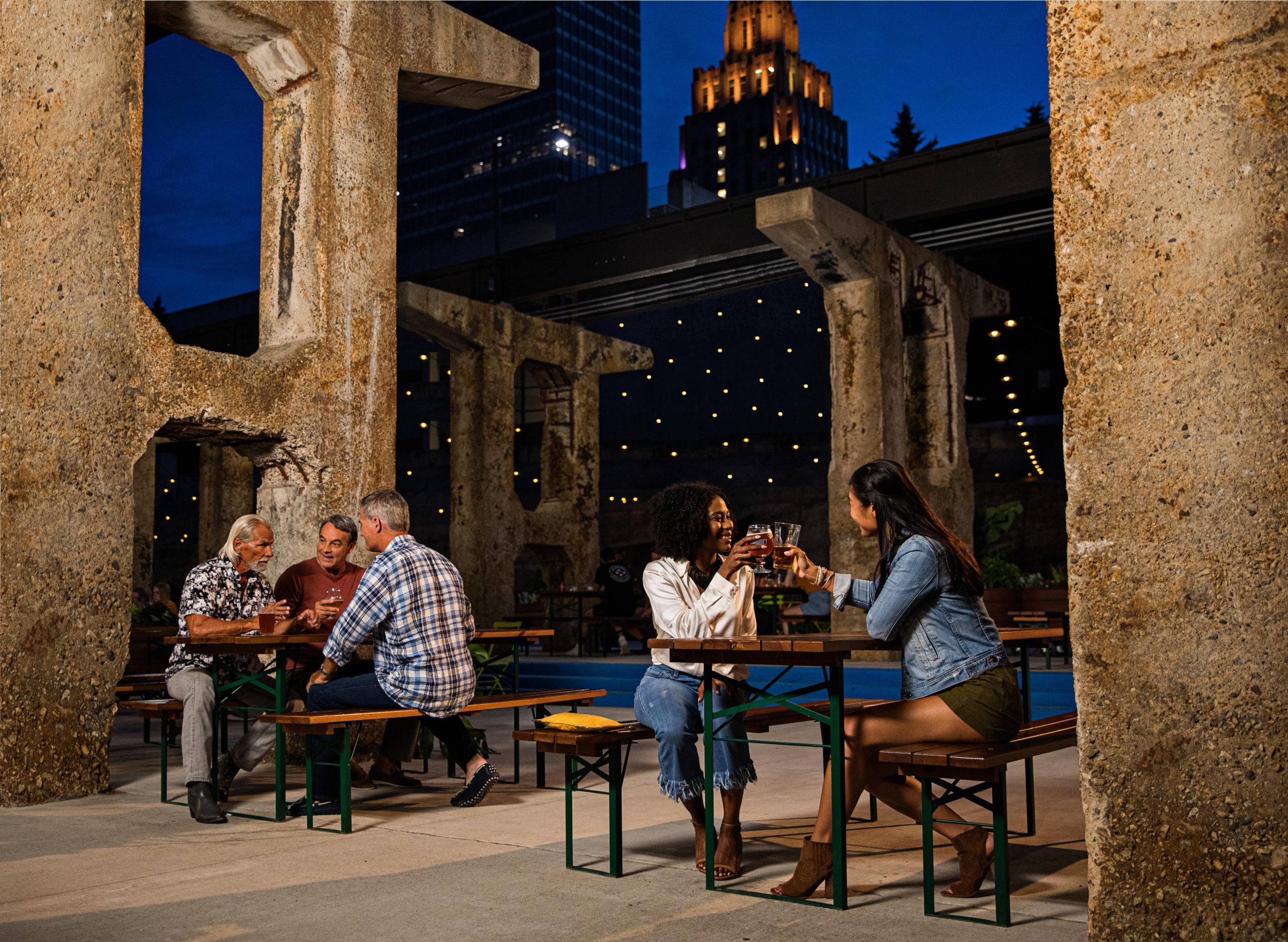 Two separate groups of people sitting in adjacent picnic tables drinking beer at a local brewery as the sun sets over the skyline above