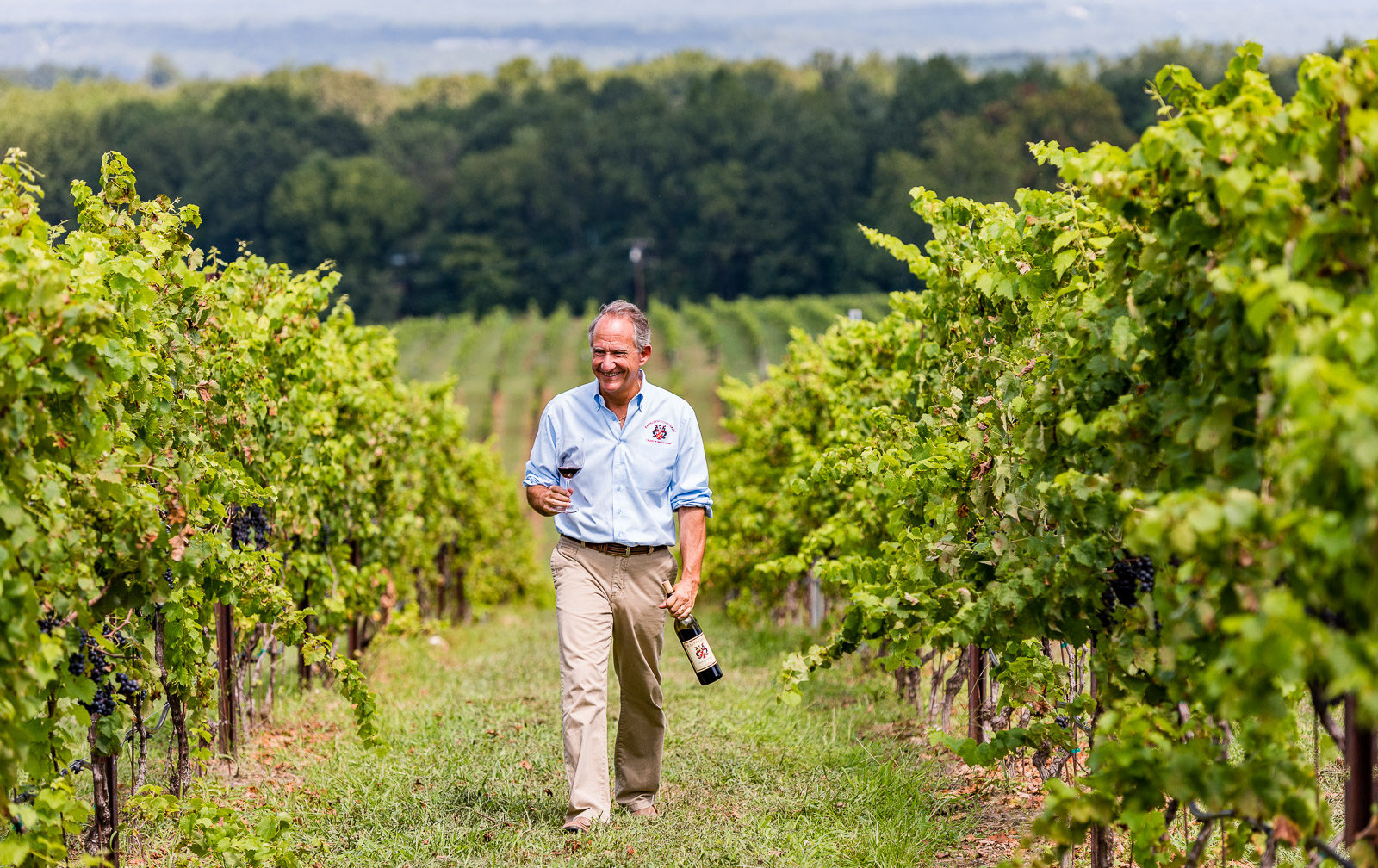 Jay Raffaldini walking the rows of vines at his namesake vineyard, holding a glass of wine in one hand and a corked bottle of wine in the other.