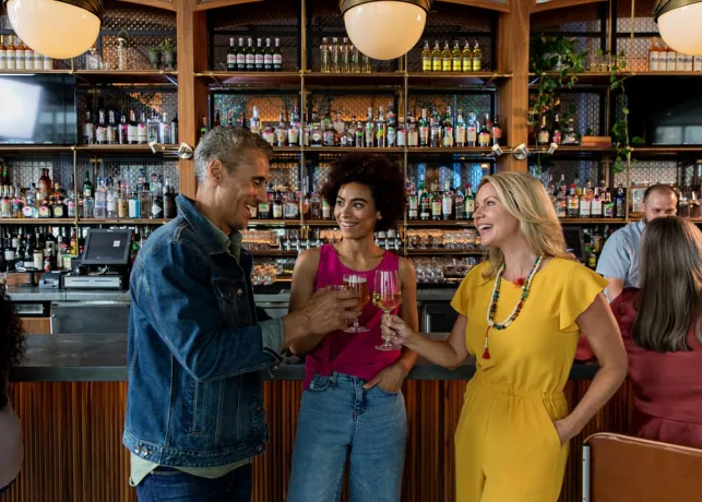 Two women and a man stand together in front of a bar clinking their glasses while chatting