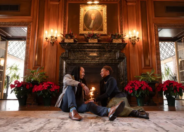 A couple sits in front of the fireplace at the Graylyn Estate, with the mantle adorned with holiday decorations.