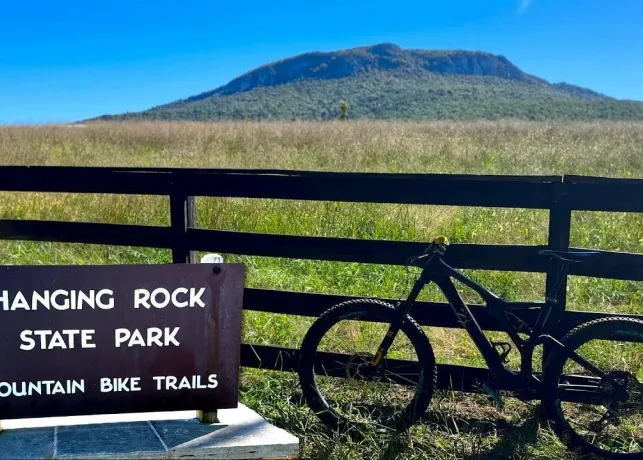 A mountain bike leans against a wooden fence next to a sign marking the entrance to Hanging Rock State Park mountain bike trails.