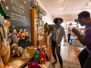 A woman stands in front of the Visual Index store signage while a man pretends to take her photo.
