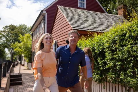A man and woman tour Old Salem Museum & Gardens together with a historic red home in the background.