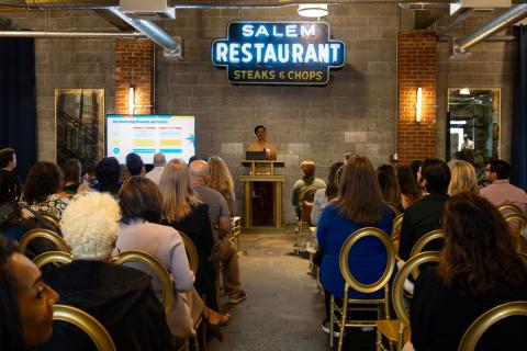 A view from the back of the crowd toward the podium while a speaker showcases a presentation on the main screen at Robert Hall Event Venue.