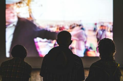 Three people sit together in the dark watching a movie screening on a large, outdoor screen.