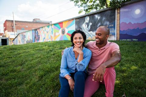 Couple sitting on grassy hill smiling, colorful mural wall behind them.