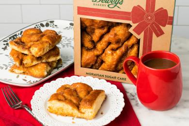 A box of Dewey's Bakery Moravian sugar cake next to a place of cake slices and a cup of coffee, sitting on top of a red place mat