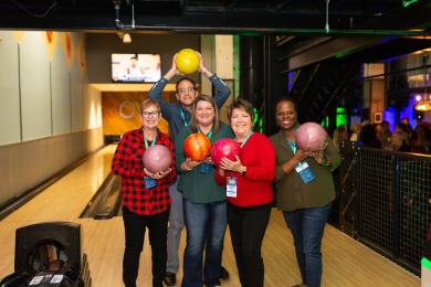 A group of five people stand together holding bowling balls before bowling in the game room at the Kimpton Cardinal Hotel.