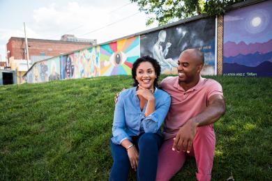 Couple sitting on grassy hill smiling, colorful mural wall behind them.