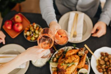Two people clinking rosé glasses over roasted chicken dinner table.