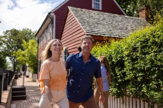 A man and woman tour Old Salem Museum &amp; Gardens together with a historic red home in the background.