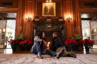 A couple sits in front of the fireplace at the Graylyn Estate, with the mantle adorned with holiday decorations.