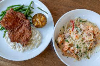 A plate of fried chicken and green beans sits on a wooden table next to another plate of shrimp and grits at Mozelle's Fresh Southern Bistro.