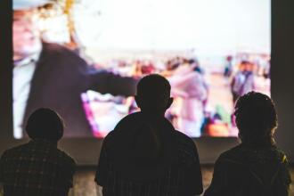 Three people sit together in the dark watching a movie screening on a large, outdoor screen.