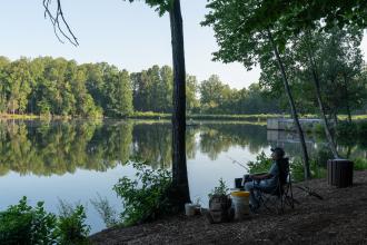 A man sits with his fishing gear in a folding camp chair near the edge of the water in Kernersville Lake Park.