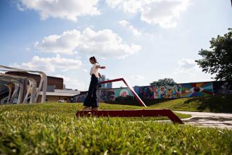 Woman balancing on red beam in grassy park with mural backdrop.