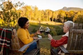 two women clinking wine glasses and sitting in wooden adirondack chairs at a winery with golden trees in the background